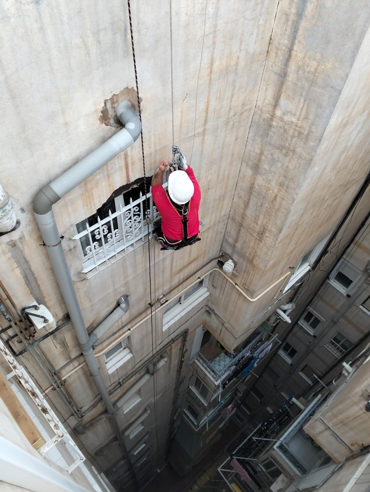 Técnico con mono rojo descendiendo por cuerda en un patio de luces junto a una bajante